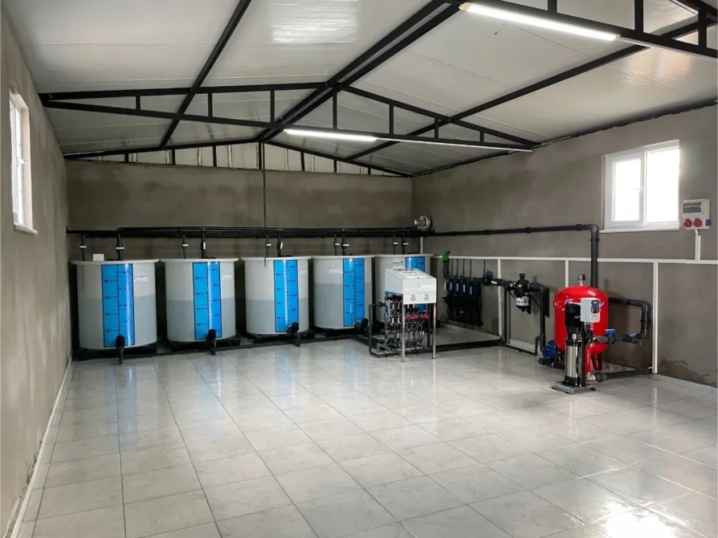 Dozaj pompası nedir?
Bir sulama sistemine ait dozaj pompası odası. Duvar boyunca sıralı beş adet mavi-beyaz karıştırma tankı, bağlantı boruları, filtreleme üniteleri ve kırmızı bir basınç tankı görülüyor. Oda temiz, fayans kaplı ve endüstriyel aydınlatmaya sahip.